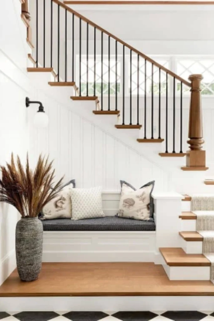 A bright, open entryway showcasing beautiful staircase landing ideas under a wooden staircase with black iron spindles. In front of a white vertical shiplap wall, a built-in bench with a dark checkered cushion and patterned throw pillows creates a cozy nook. On the light wood floor platform next to the bench, a tall, artful grey textured stone pot holds a large arrangement of dried pampas grass.