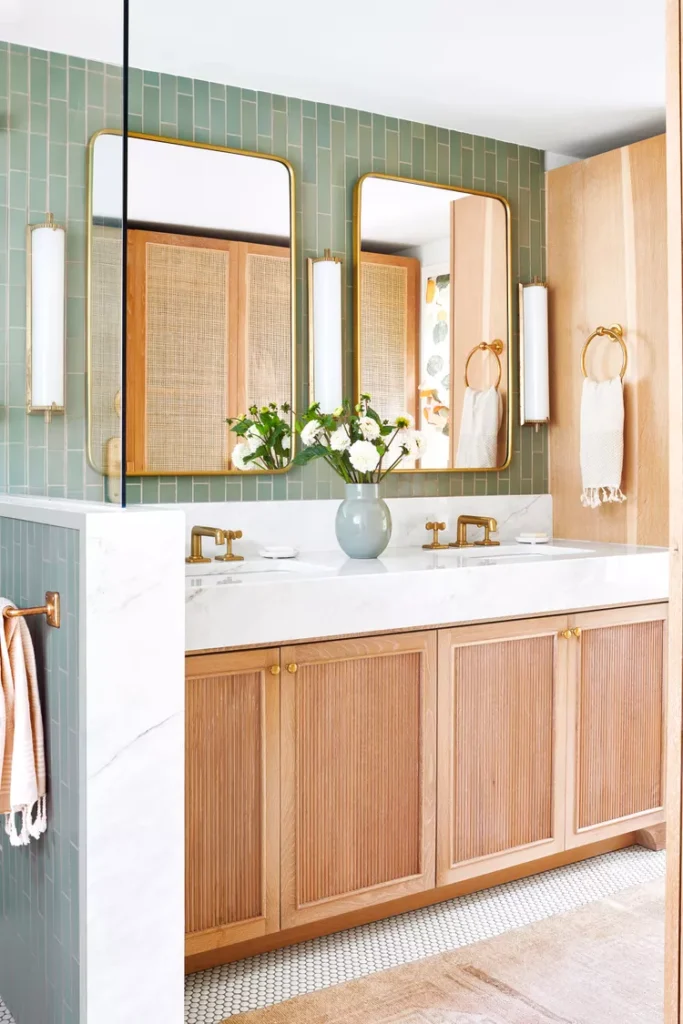 A luxurious master bathroom vanity showcasing the best bathroom ideas master bath design trend. The space features a sage green vertical subway tile accent wall behind twin gold-rimmed rectangular mirrors and brass fixtures. A warm fluted wooden vanity with a white marble countertop is beautifully complemented by matching cane-insert wardrobe doors reflected in the background.