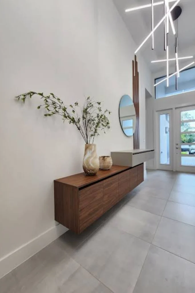 A modern minimalist hallway featuring a dark wood floating console unit mounted on a white wall, styled with a tall ceramic vase with greenery and a round mirror, demonstrating sleek Apartment Rooms ideas for a functional drop zone.