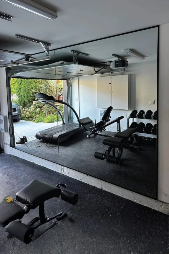 A functional garage home gym with a large, full-length mirror wall on concrete flooring, reflecting a treadmill, weight benches, and a rack of dumbbells next to an open garage door looking out onto a garden.