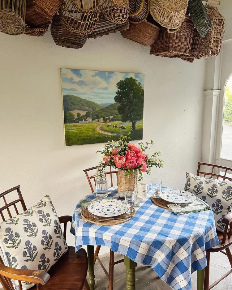 A rustic dining setup showcasing small screened-in porch ideas decor, featuring a round table with a blue checkered tablecloth, floral patterned cushions on wooden chairs, a large countryside landscape painting on the wall, and a unique display of woven baskets hanging from the ceiling.