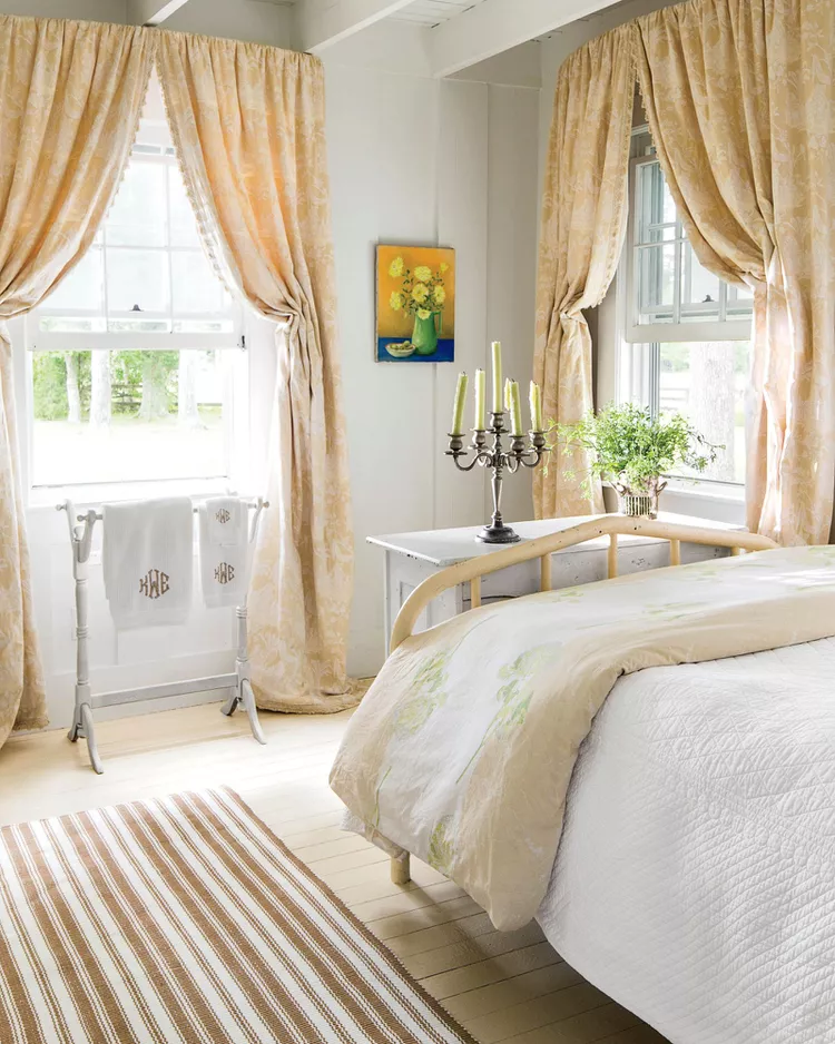 A bright country-style bedroom layout demonstrating how to use bedroom flooring ideas like painting a floor with a fresh coat to brighten a dark room. The space features light cream painted wood plank flooring accented with a brown and white striped area rug. A vintage iron bed frame sits between two large windows framed by long, flowing beige floral curtains, under high ceilings with open white beams.