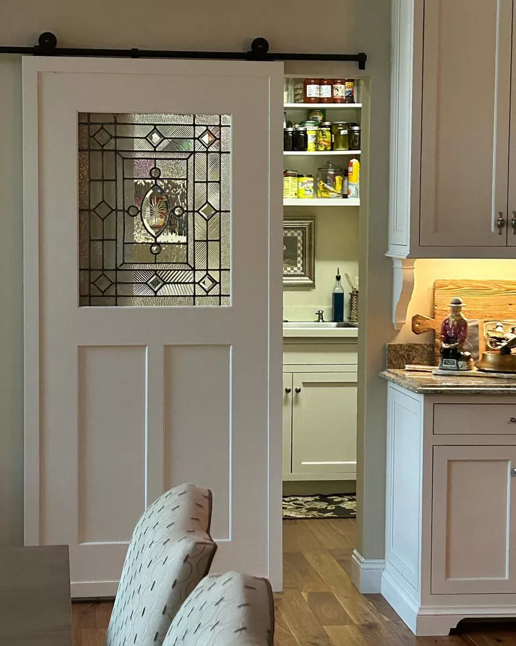 A white sliding barn door featuring a stunning, geometric leaded stained glass window panel, showcasing how to give a sliding door new life by using texturized glass inserts to separate a kitchen from a pantry area.