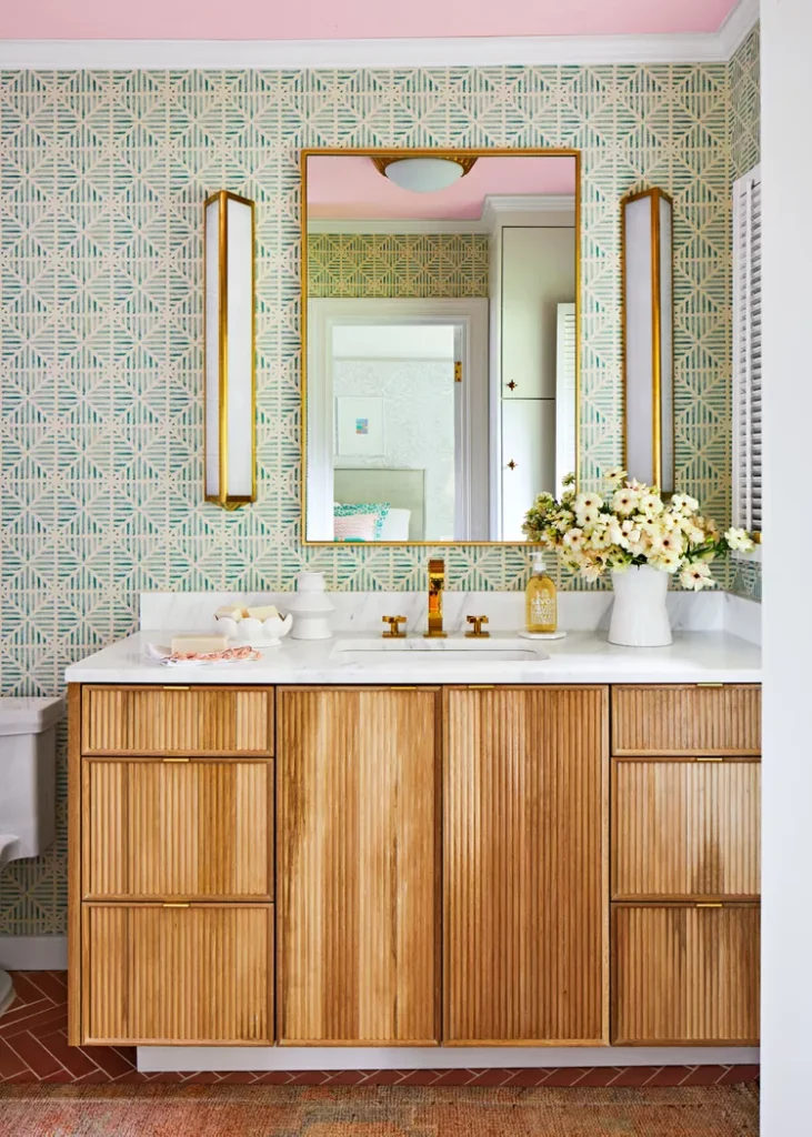 A beautifully styled bathroom vanity illustrating how to go all in on earthy textures for a warm, organic feel. The layout features a rich, fluted light-wood vanity cabinet with a clean white marble countertop and brass fixtures. The background accent wall is covered in a textured green and cream geometric block-print wallpaper, perfectly framing a gold rectangular mirror and two long architectural brass wall sconces.