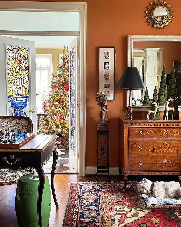 An elegant room interior with warm terracotta walls, an antique chest of drawers, and an open doorway featuring a highly intricate, colorful floral stained glass window panel that brings a vibrant Mediterranean jewel-toned design into a traditional living space.