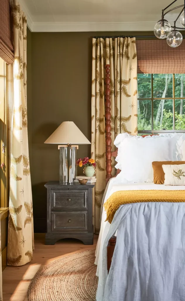 An organic modern biophilic bedroom layout showcasing beautiful bedroom flooring ideas by choosing to go natural underfoot. The space features solid hardwood flooring in a light warm oak tone, partially covered by a circular braided jute area rug next to an antique four-poster wooden bed frame. The room is styled with earthy olive green walls, a dark wood nightstand, and cream curtains featuring a brown fern leaf print pattern.