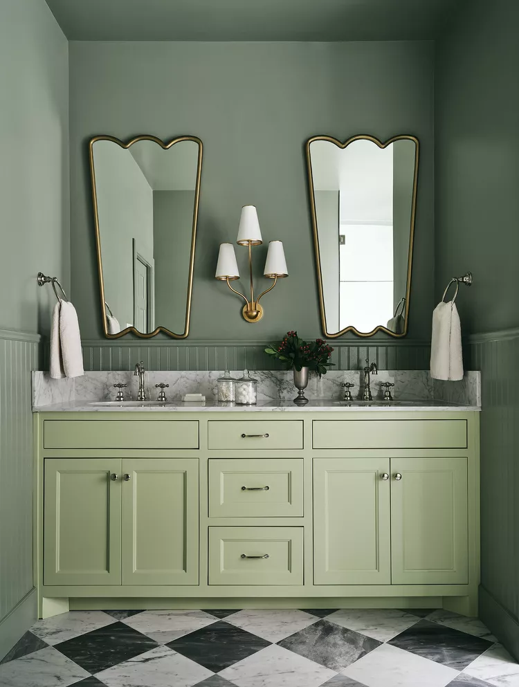 A sophisticated green bathroom vanity illustrating a smart two-toned alternative to the gray best bathroom ideas modern designers often use. The space features a large olive green double vanity with a white marble countertop, set against sage green walls and matching beadboard wainscoting. Two gold wavy-edged mirrors and a central brass multi-shade wall sconce accentuate the layout above a striking black and white checkerboard marble tile floor.