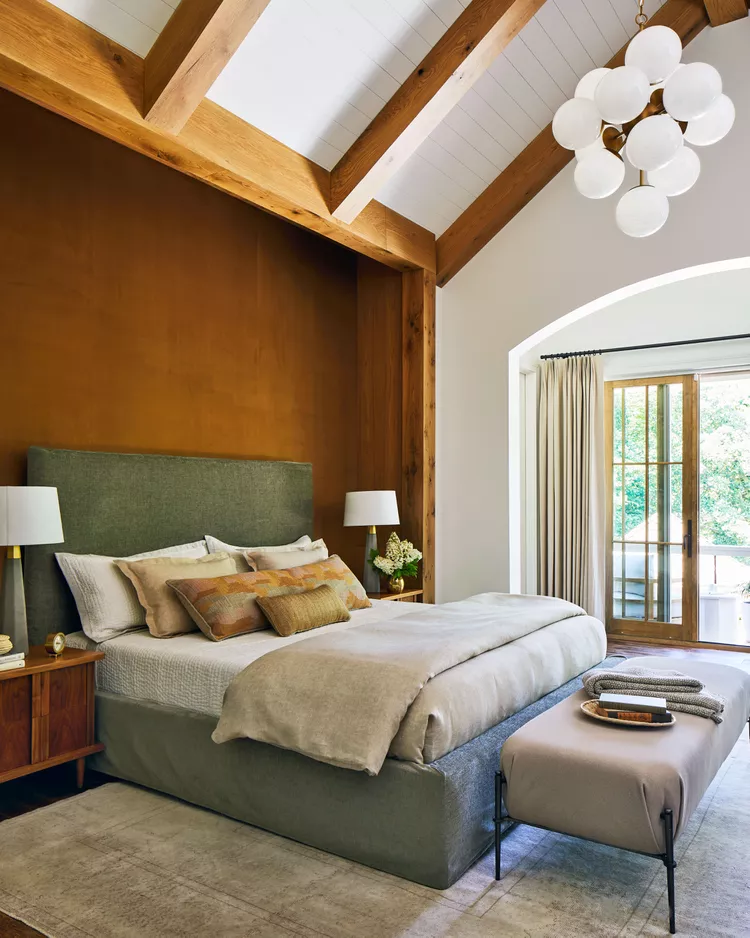 A rustic modern master bedroom highlighting authentic bedroom flooring ideas by choosing to go vintage with historic and distressed materials. The layout features rich, dark distressed hardwood flooring partially covered by a large, faded neutral vintage oriental rug beneath a gray upholstered platform bed. The room boasts a striking warm terracotta accent wall behind the bed, complemented by high vaulted ceilings with massive exposed natural wood beams and a modern white globe cluster chandelier.