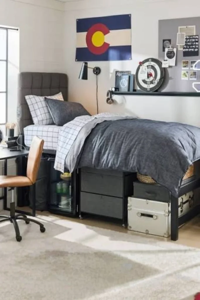A modern and cozy college room featuring stylish gray bedding dorm room ideas, with a lofted twin bed, a charcoal gray comforter layered over grid-patterned sheets, under-bed storage drawers, a small fridge, and a brown leather desk chair in a sunlit room.