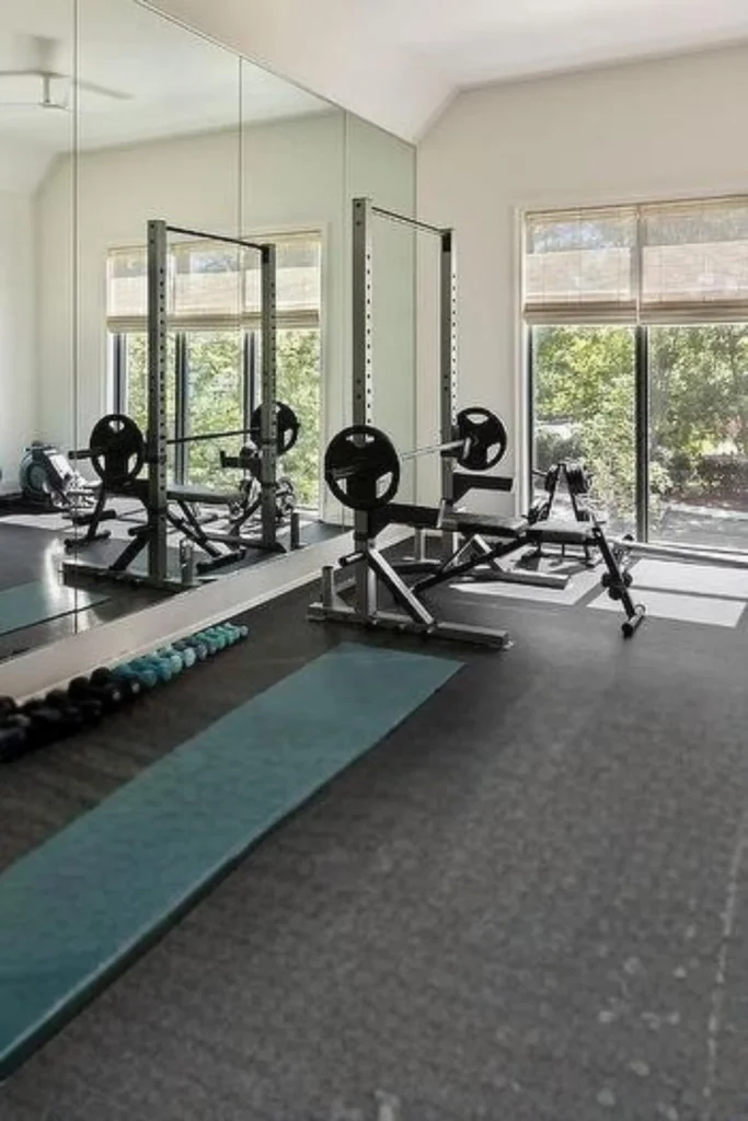 A modern home gym setup featuring a large grid glass panel mirror wall mounted behind a weight bench, barbell rack, and dumbbells, reflecting natural light from an adjacent window.