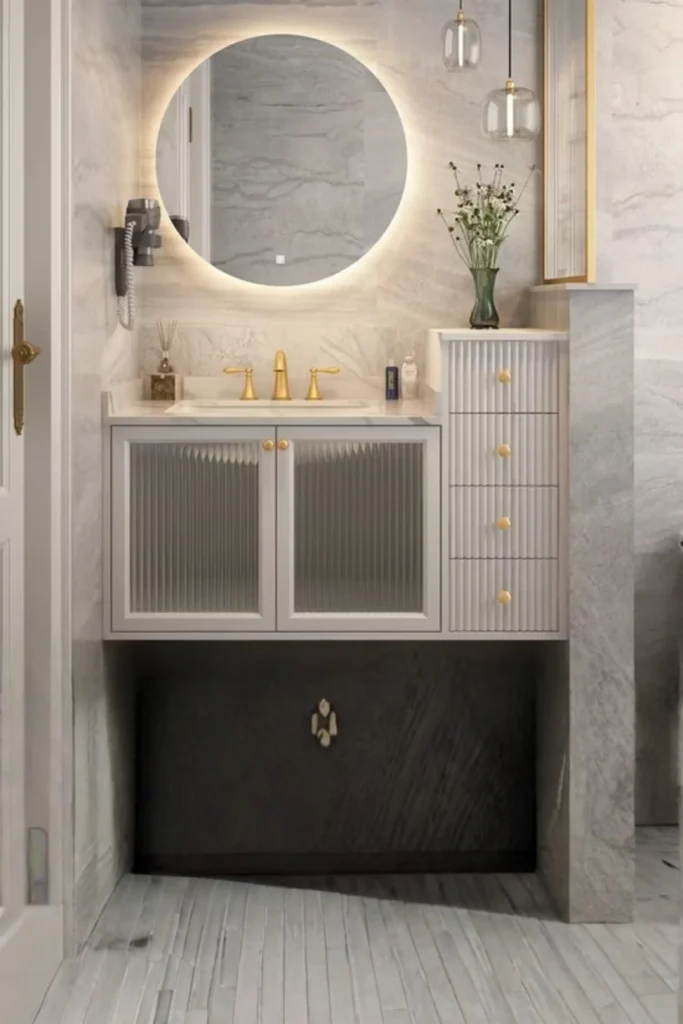 A luxury modern bathroom vanity layout highlighting how hanging a new, uniquely shaped mirror instantly alters the visual flow and adds a heavy dose of luxury style. The space features a large round backlit LED mirror mounted over a white fluted floating vanity cabinet with ribbed glass doors and elegant brass fixtures, all set against beautiful gray marble-textured walls and flooring.