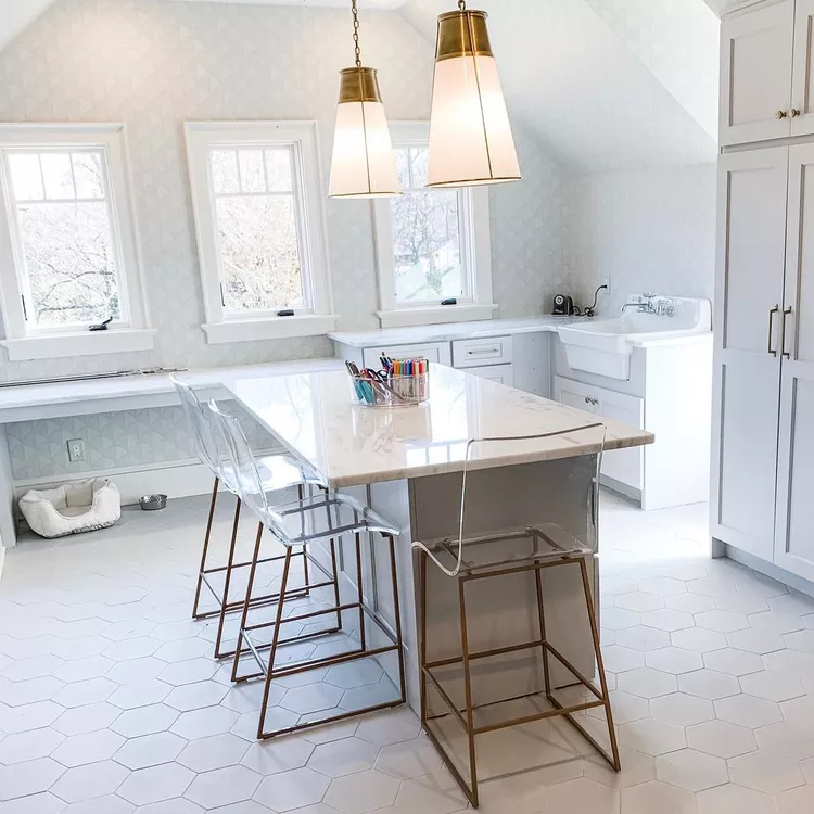 A bright, all-white attic studio room featuring white hexagonal tile flooring and soft geometric patterned wallpaper on the vertical walls. Hanging above a central marble-topped island with clear acrylic barstools are two large brass cone pendant lights. The layout showcases custom white cabinets, a large farmhouse apron sink, and three symmetrical windows set beneath a deeply pitched roofline, demonstrating beautiful ways to highlight the slanted ceiling as a prominent architectural feature in a renovated attic space.