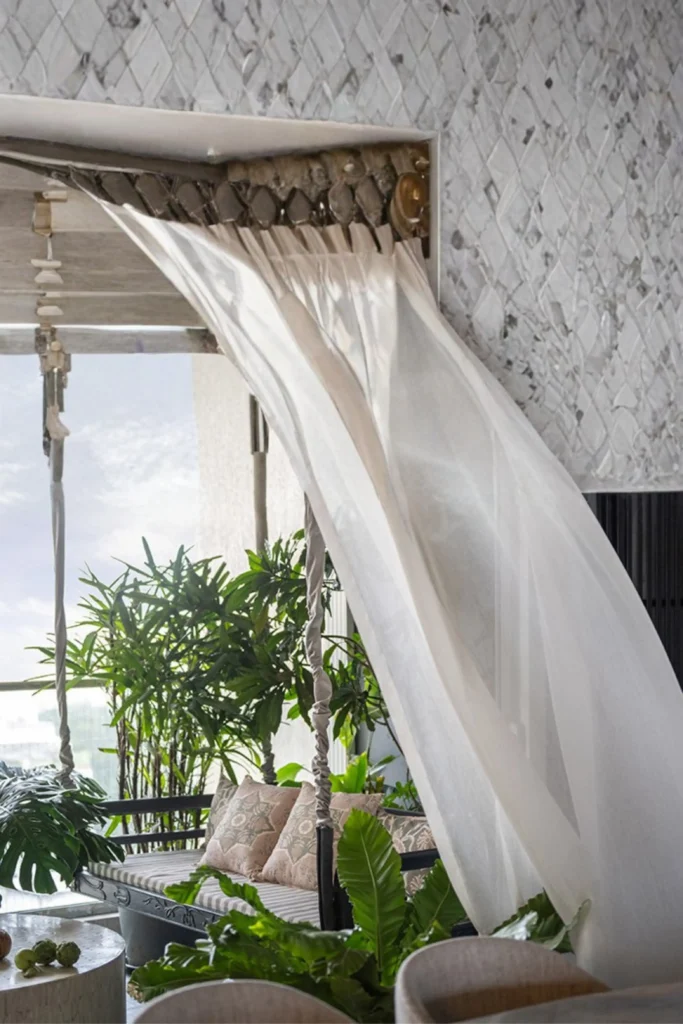 A breezy balcony terrace showing how to incorporate lush, beachy greenery into your home layout. A sheer white curtain blows in the wind next to a textured mosaic tile wall, revealing an outdoor daybed styled with patterned neutral pillows. The space is surrounded by vibrant, tropical palm trees and broad-leaf monstera plants that bring a refreshing beachside texture to the scene.