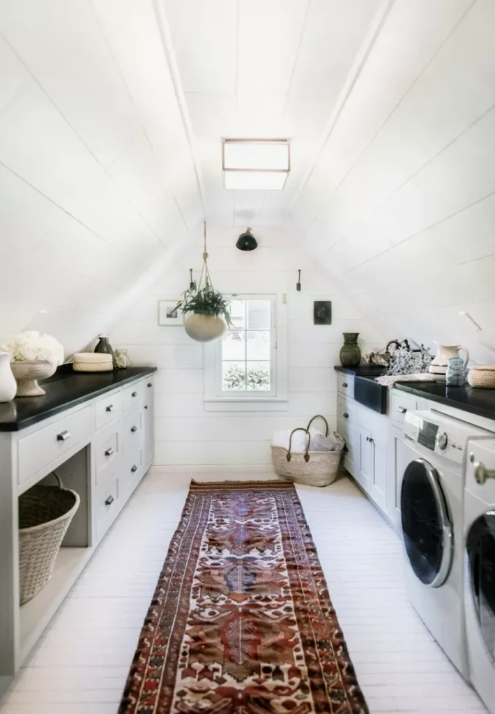 A long, narrow attic space transformed into a bright laundry and storage room, featuring a patterned vintage runner rug on white wood floors. The walls and deeply pitched, sloped ceilings are lined with clean white shiplap paneling. Custom built-in cabinets with dark countertops run flush along the low eaves on both sides, integrating a washing machine, dryer, and storage cubbies, perfectly illustrating how attic closet ideas angled ceilings diy projects can maximize a pitched roof with tailored cabinetry.