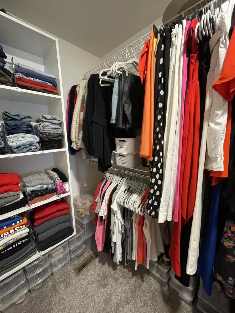 An organized walk-in closet space featuring a white built-in shelving tower filled with neatly folded t-shirts, pants, and clear plastic shoe storage bins along the floor. Maximize your vertical height by installing a double-rod system with wire shelving, showcasing rows of colorful shirts, blouses, and dresses hung neatly on matching white hangers, demonstrating practical wardrobe organization tips for small spaces.