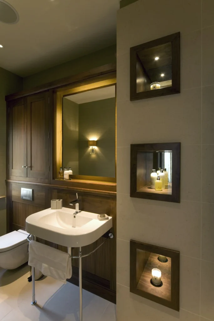 A sophisticated, modern bathroom showcasing smart bathroom remodel ideas for tight floor plans. The space demonstrates how installing a custom recessed wall niche maximizes vertical real estate, offering a streamlined, professional alternative to bulky storage. The design features a white pedestal sink, rich wood paneling, and three illuminated, custom-built wall niches set into a neutral tiled wall, perfectly displaying skincare products and candles.