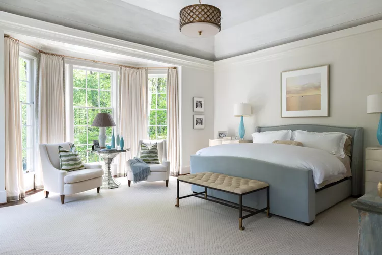 A spacious luxury master bedroom displaying premium bedroom flooring ideas by investing in custom, high-end floor coverings. The layout features a seamless, white low-profile textured patterned carpet running wall-to-wall under a contemporary curved gray bed frame. The room is styled with white walls, a large bay window area with cream drapes, twin accent armchairs, and a stylish brass drum pendant light hanging from a high tray ceiling.