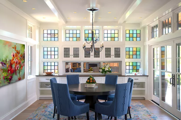 A luxury dining room layout with a round table and blue chairs, surrounded by built-in white cabinetry and custom gridded glass window panes featuring subtle colored inserts to jazz up the dining room and flood the space with natural light.