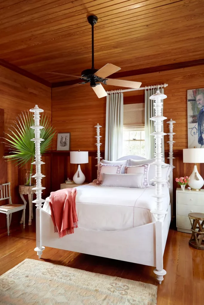 A warm, wooden-themed master bedroom displaying elegant bedroom flooring ideas by keeping the layout minimal and clean underfoot. The space features highly polished solid wood plank flooring running seamlessly under a unique white spindle four-poster bed. A simple, neutral low-pile vintage area rug anchors the foreground, while matching warm horizontal wood plank panels wrap the walls and ceiling for a cozy cabin aesthetic.