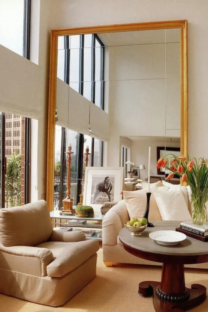 A spacious, sunlit living room featuring Large Floor Mirrors with a gold frame leaning against a high wall, reflecting large windows and beige seating to double the natural light.