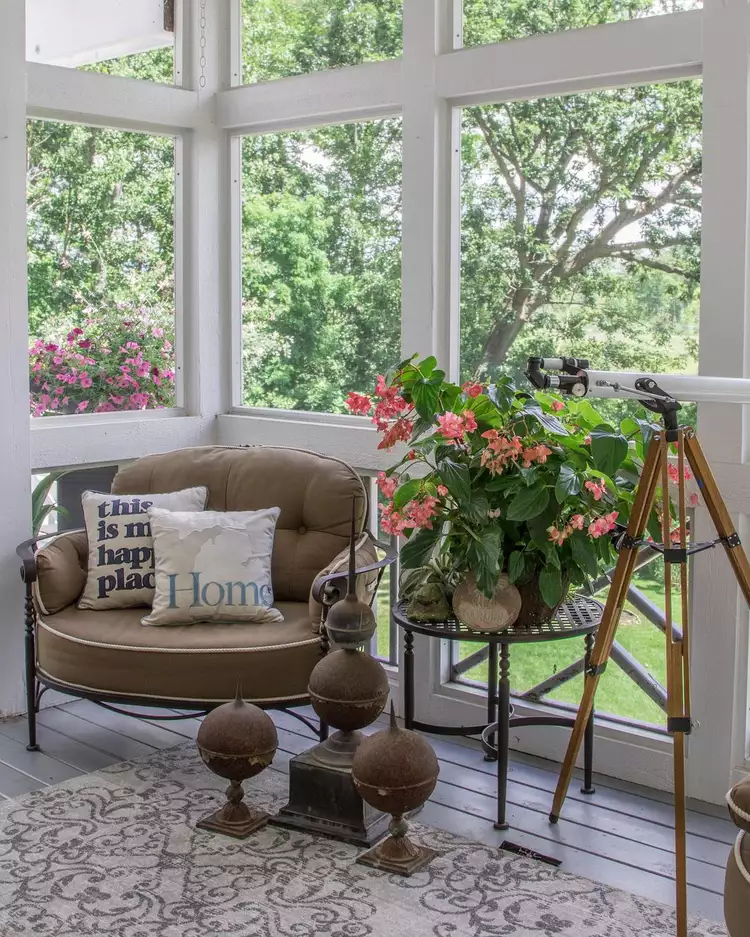 A cozy setup showcasing small screened-in porch ideas decor, featuring a grey patterned outdoor rug laid on a wooden floor, anchoring a comfortable brown armchair with throw pillows, a plant stand, and a vintage tripod telescope by the window.
