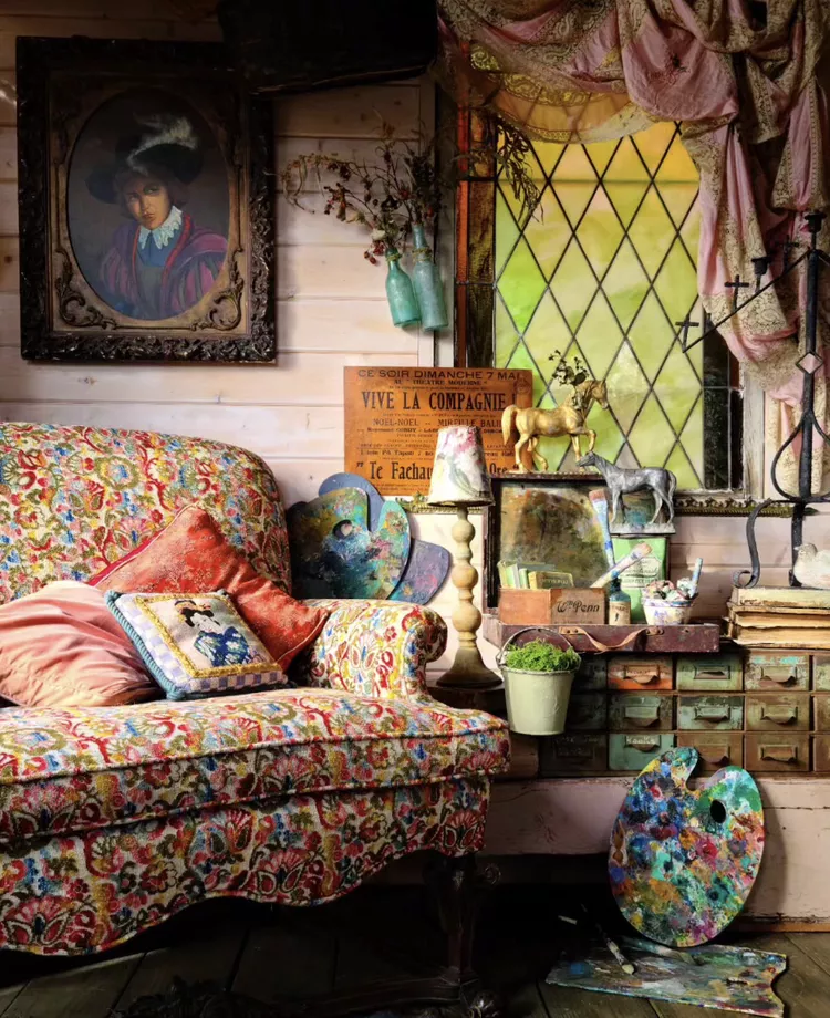 A cozy, artistic reading nook featuring a vintage floral sofa and an antique dresser assembly beneath a classic diamond-paned leaded glass window, highlighting cozy glass window ideas to add warmth and character to a private room layout.
