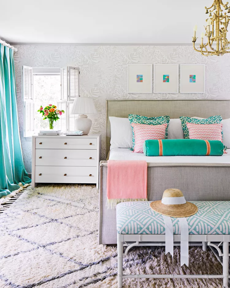 A bright, luxurious master bedroom showcasing cozy bedroom flooring ideas by making the space ultra-plush for ultimate comfort. The layout features an ultra-thick, shaggy white Moroccan-style wool rug with black diamond lines beneath a tufted gray upholstered bed frame. In the foreground, another dense, high-pile textured gray shag rug rests under a patterned light-blue accent bench, beautifully matching teal curtains and a subtle white leaf-print wallpaper wall.
