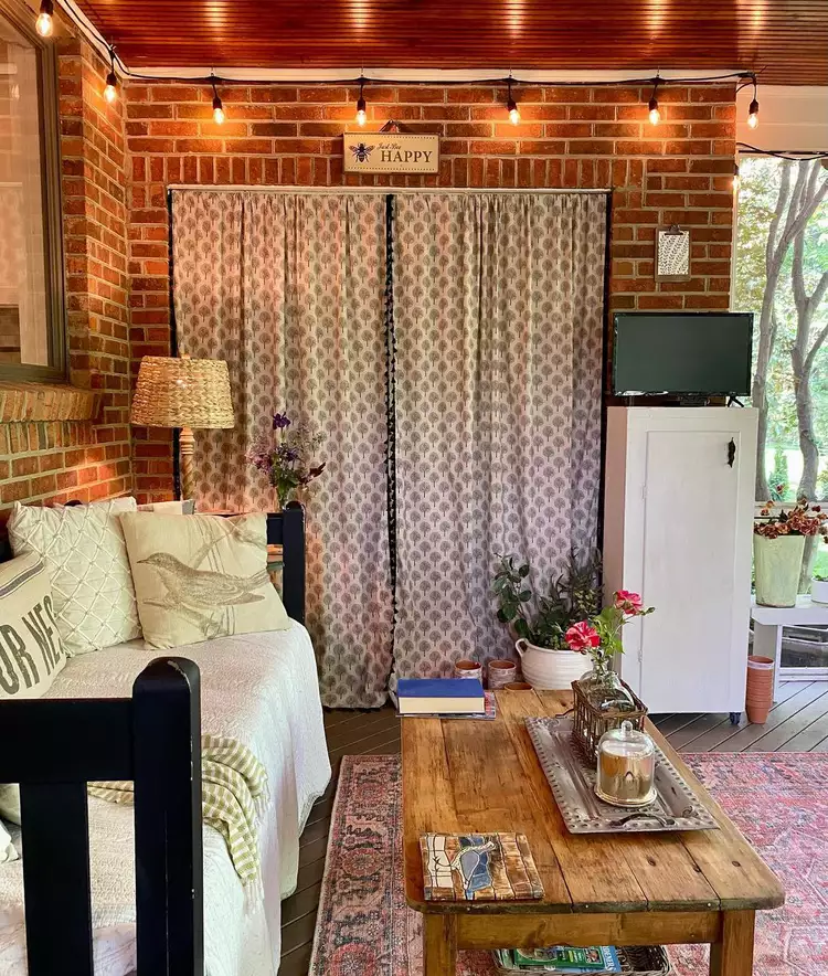 A cozy seating arrangement showcasing small screened-in porch ideas, featuring a long rustic wooden coffee table decorated with a tray and flowers, set against an exposed brick wall, string lights, and a comfortable daybed with throw pillows.