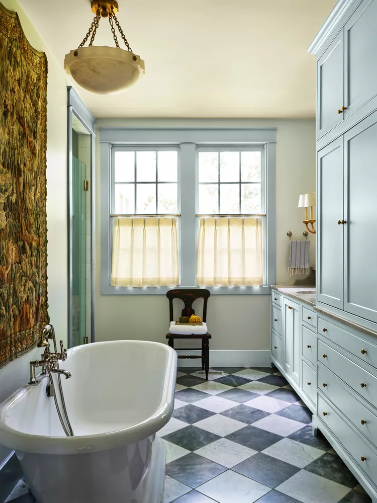 A classic bathroom highlighting one of the best bathroom ideas to elevate a room with bold flooring. The space features a striking black and white checkerboard diamond marble tile floor, which perfectly anchors a freestanding white clawfoot soaking tub and large pale blue built-in vanity cabinets. Light enters through double windows with soft cream tier curtains, under an elegant vintage pendant light fixture.