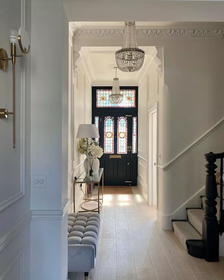 An elegant hallway view leading to a black front door under tall ceilings, featuring brilliant geometric stained glass windows and a transom pane that maximize the home's scale and light.