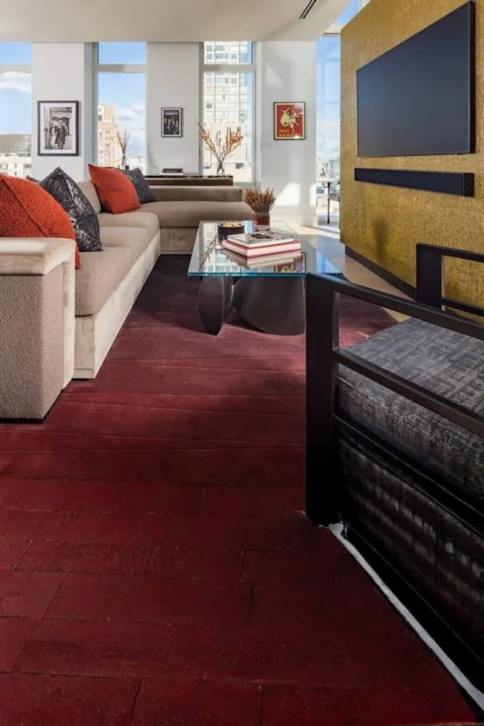 A modern high-rise living room showcasing how to mix different tactile elements, perfectly matching contemporary and modern house interior decorating ideas bedroom aesthetics. Features a large neutral sectional sofa styled with vibrant burnt orange pillows, a sleek glass coffee table with a metal base, and a deep reddish-brown textured area rug set against an accent wall holding a wall-mounted TV.