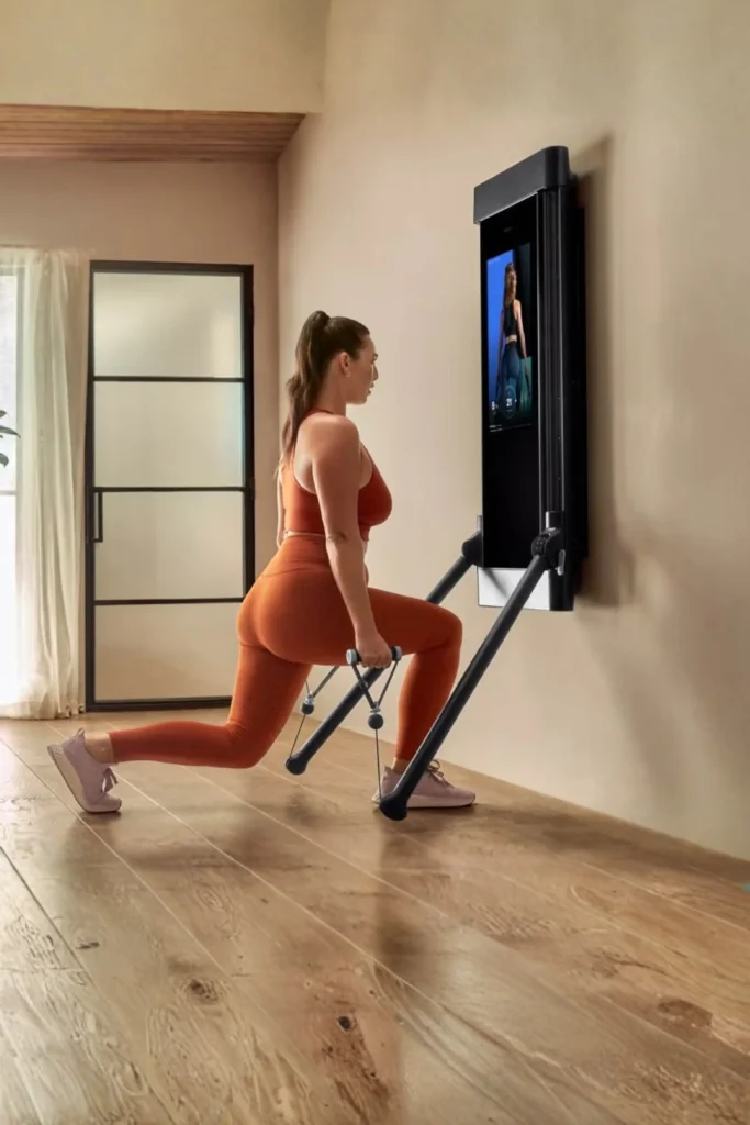 A woman in rust-orange athletic wear performing lunges using resistance cables attached to a sleek, wall-mounted smart gym display mirror in a modern, sunlit room with wooden floors.
