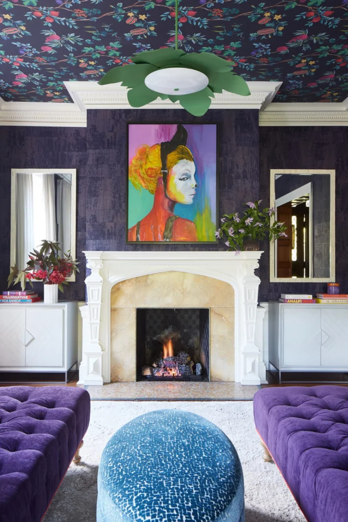 A luxurious living room embodying the Moody Elegance: Deep, Enveloping Tones design trend. The space features a dark purple textured wallpaper accent wall surrounding a classic white fireplace mantel with a vibrant colorful portrait hanging above it. The ceiling is uniquely styled with a dark floral wallpaper pattern, complemented by rich purple tufted velvet sofas and a blue textured oval ottoman on a plush white rug.