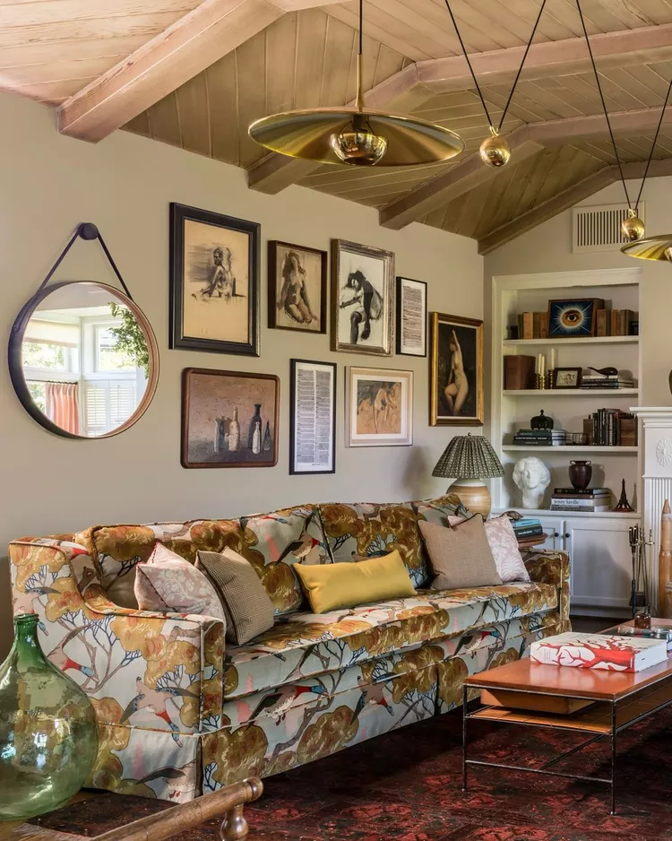 A warm, eclectic living room with a colorful patterned sofa and a gallery art wall, featuring a beautiful vaulted sloped ceiling lined with warm natural wooden planks and exposed beams to bring organic texture and a comforting, rustic sanctuary feel to the space.