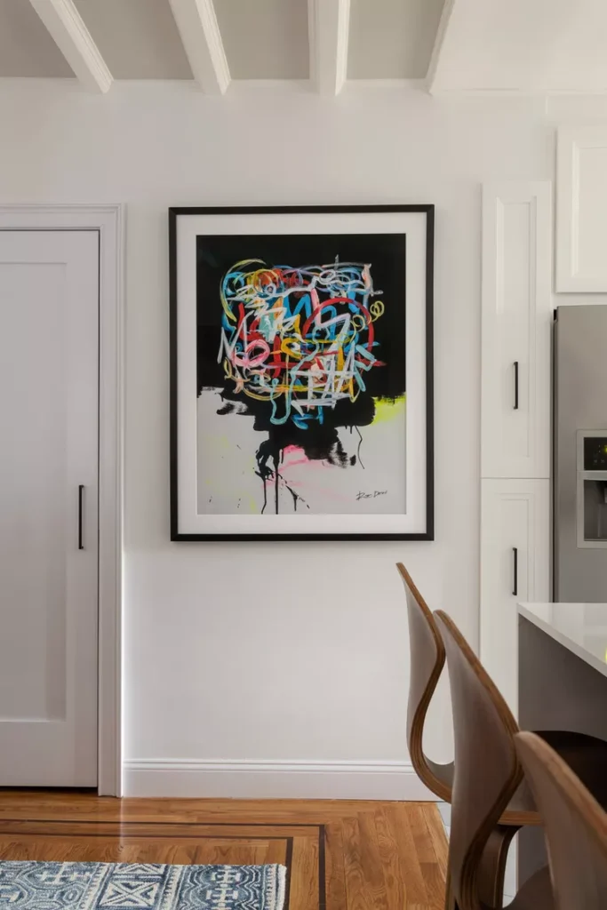 A bright, modern kitchen and dining space with white walls, warm hardwood floors, and a large, vibrant abstract artwork framed on the wall, balanced beautifully by a soft, warm gray neutral ceiling featuring simple white structural beams to show how keeping your ceiling softly neutral creates a spacious, calm, and elegant room.