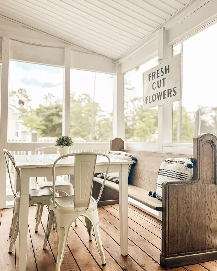 A farmhouse-style setup for small screened-in porch ideas, featuring a long vintage wooden dining bench with boho throw pillows paired with a white wooden dining table and metal chairs on a wood plank floor.