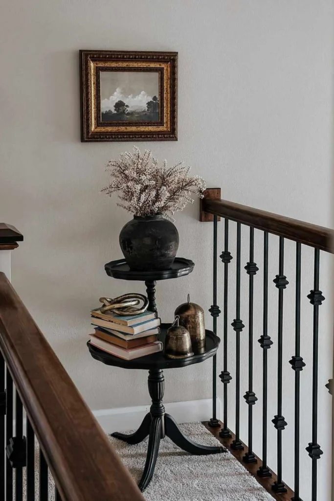 A close-up of a traditional stairwell landing showcasing clever staircase landing ideas for small spaces. A unique black wooden two-tiered pedestal table sits perfectly in the corner next to a dark wood handrail with black iron balusters. The top tier holds a rustic black vase with dried white flowers, while the lower tier is styled with a stack of antique books, a gold knot sculpture, and vintage brass bells under a small framed landscape oil painting on a neutral wall.