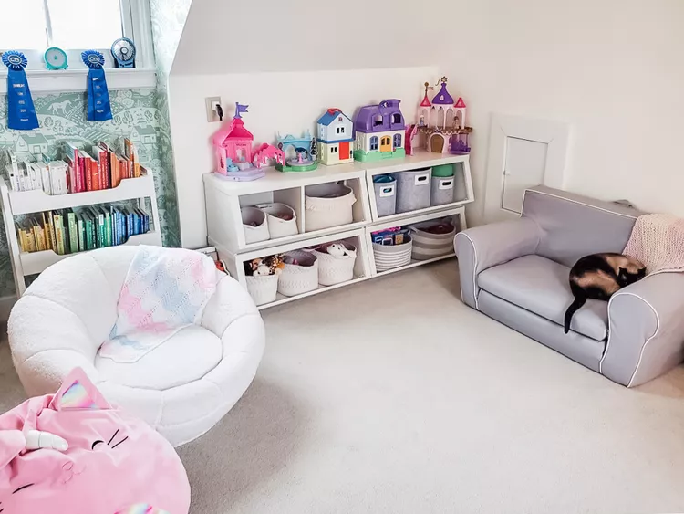 A cozy child's playroom and multi-purpose family zone arranged beneath a low sloped attic wall. The setup features a small gray child-sized sofa, a white round sherpa chair, and a white tiered bookshelf filled with books. Arranged neatly along the floor directly beneath the angled pitch are stacked low open cubbies filled with soft fabric storage bins and plastic tubs holding toys, with dollhouses displayed on top, showing a brilliant way to organize toys using low eave sections.