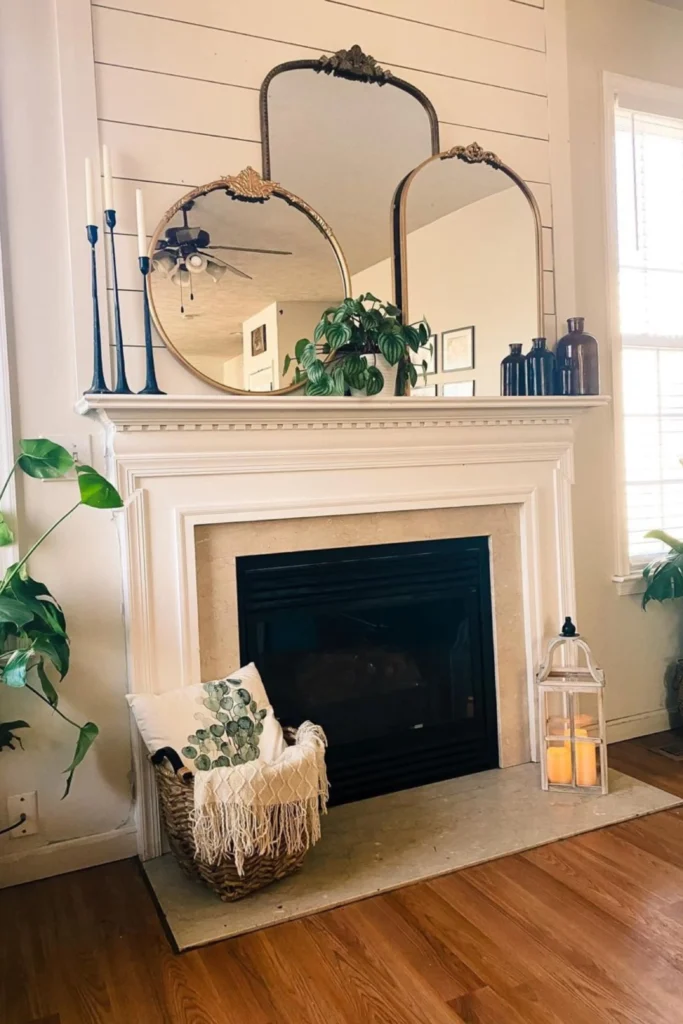 A cozy white shiplap fireplace mantle decorated with layered Oversized Mantle Mirrors featuring vintage gold frames, styled with slender black candlestick holders and potted green house plants.