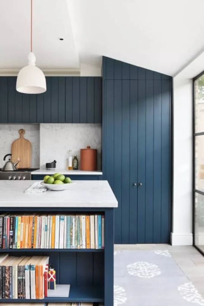 A modern kitchen displaying clever navy blue accent wall ideas by using floor-to-ceiling shiplap paneled cabinets in deep navy. In the foreground, a matching navy kitchen island features built-in open bookshelves packed with colorful cookbooks under a white marble countertop, accented by a white dome pendant light and light gray patterned floor runner.