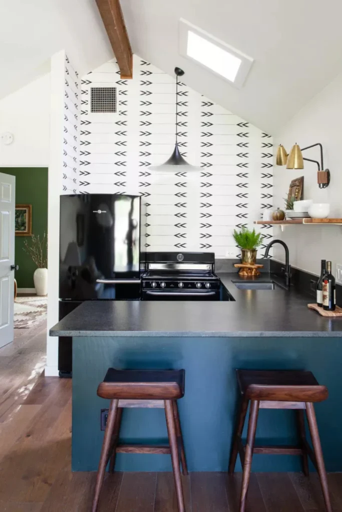 A cozy, modern kitchen showcasing unique navy blue accent wall ideas by painting the backside of the lower breakfast bar cabinets in deep navy. This design creatively experiments with navy blue and grey accent wall ideas, paired with sleek dark wooden bar stools, black quartz countertops, an industrial black pendant light, and a striking black and white patterned arrow accent wall under a sloped ceiling with a skylight.