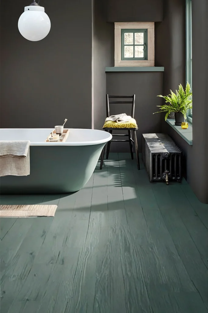 A moody and sophisticated space featuring high-end modern Bathroom Remodel Ideas. The bathroom showcases deep charcoal gray painted walls that contrast beautifully with a muted slate-green freestanding bathtub and matching painted wood plank flooring. Natural light streams in from a window ledge decorated with a vibrant green fern in a black pot, creating an intimate, cozy spa atmosphere.