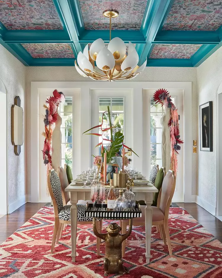 An eclectic, high-end dining room with a patterned pink area rug, a fully set dining table, and a modern white-and-gold lotus chandelier, crowned by a painted coffered ceiling with wallpaper featuring teal blue painted grid lines that frame a colorful, detailed floral wallpaper inside the recessed panels to showcase how combining woodwork, paint, and patterns creates a custom designer ceiling.