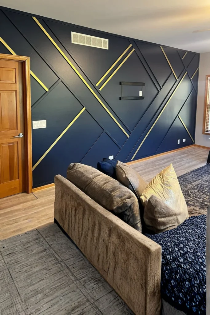A luxury living room showcasing bold navy blue accent wall ideas with a custom geometric wood trim design. The accent wall features intersecting dark blue panels accented with striking polished gold metallic stripes. In the foreground, a comfortable beige velvet sofa decorated with gold and navy blue cushions sits on a textured rug over light wooden flooring.