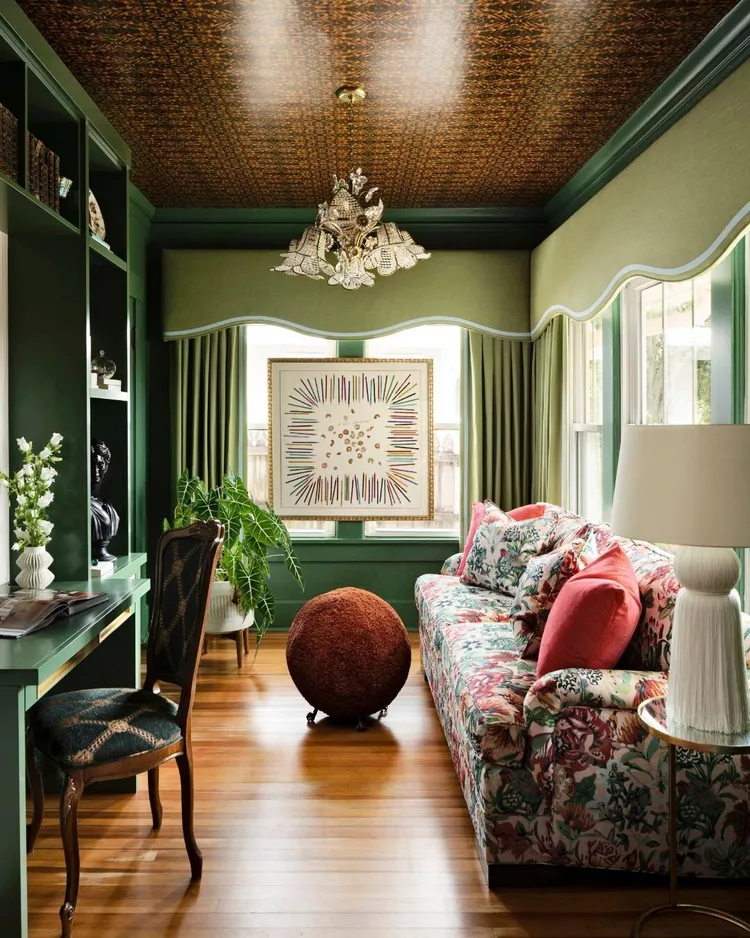 A vibrant, cozy sitting room with deep green walls, custom built-in shelves, and a colorful floral print sofa, crowned by a peppy patterned wallpaper in a detailed brown geometric design on the ceiling to show how unexpected patterns overhead add designer flair and joy.