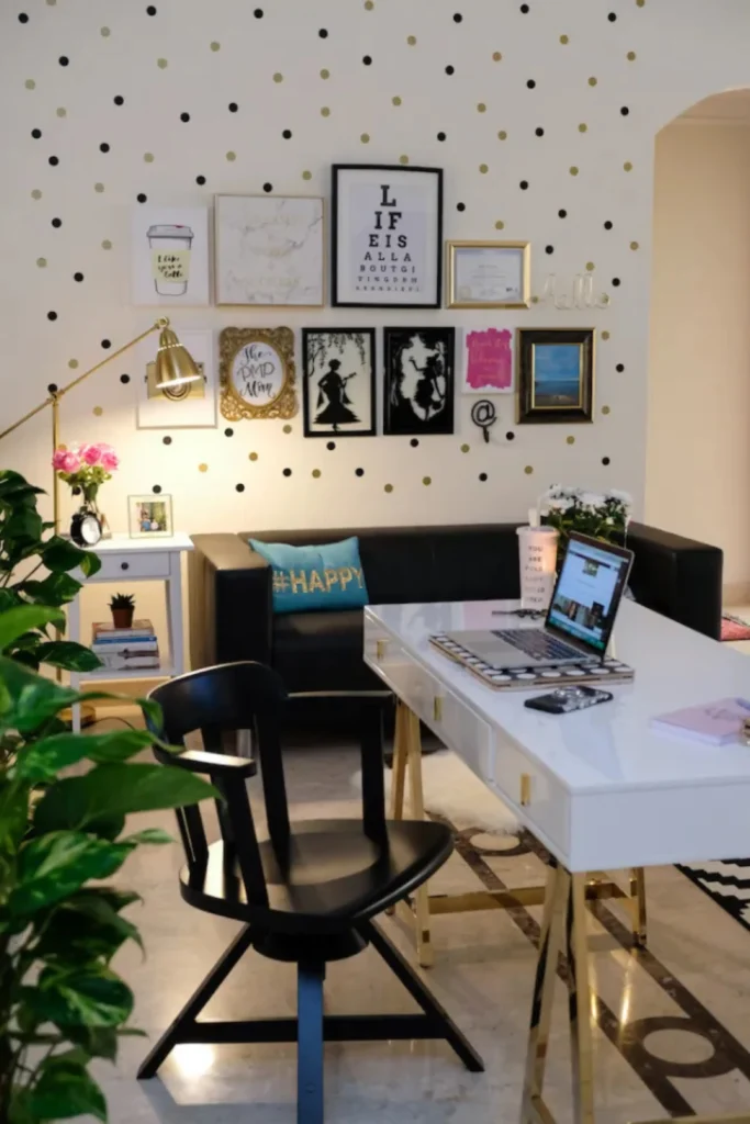 A stylish home office featuring a personalized gallery wall displayed on polka dot wallpaper, styled with framed typography prints, inspirational art, and certificates above a black leather sofa and a glossy white desk with gold legs.