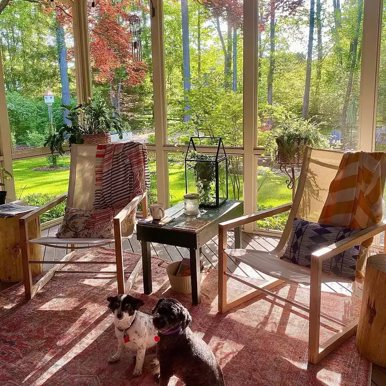 A practical outdoor setup showcasing small screened-in porch ideas decor, featuring two modern wooden rocking chairs with performance mesh fabric, outdoor throw blankets, a red patterned rug, and a narrow black wooden side table.