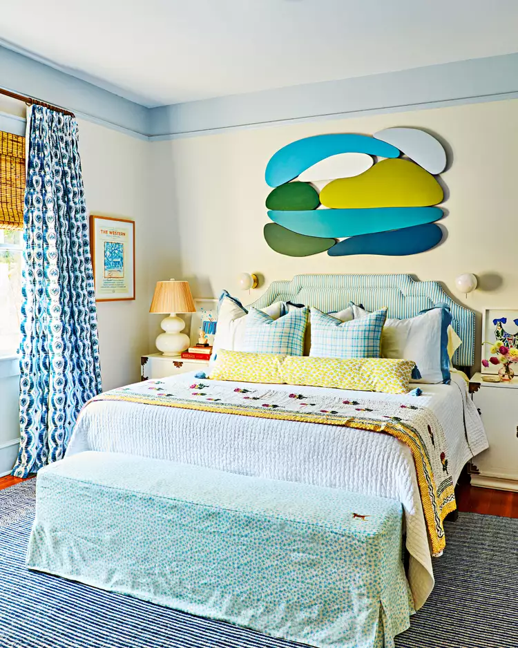 A vibrant, eclectic bedroom layout demonstrating creative bedroom flooring ideas by using a bold patterned rug over a rich hardwood floor to act as a work of art. The space features a classic dark wood floor layered with a dense navy and white striped area rug under a bed with a light blue striped headboard. The room is styled with colorful abstract organic-shaped wall art, patterned blue curtains, and a speckled blue fabric bench at the foot of the bed.