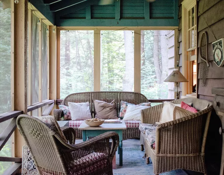 An indoor-outdoor transition showcasing small screened-in porch ideas cozy styling, featuring a full wicker living room set with soft throw pillows, a central wooden coffee table, an accent lamp, and subtle string lights under a dark teal ceiling.