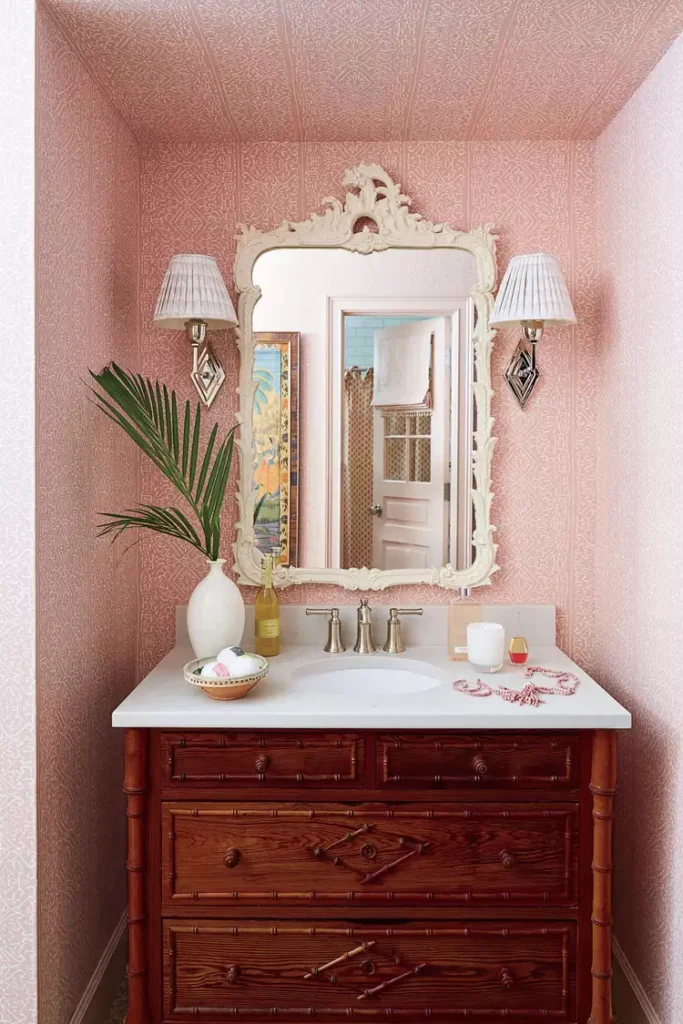 A charming powder room showcasing one of the best bathroom ideas small spaces require for a cozy vanity setup. The image features a vintage dark wood chest converted into a washbasin vanity with a white countertop. The alcove walls and ceiling are wrapped in a soft pink patterned wallpaper, framing an elegant white ornate mirror and two classic pleated fabric wall sconces.